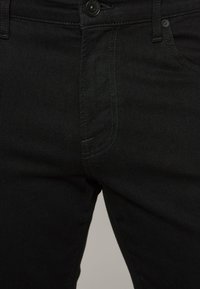 I jeans in denim neri presentano una vestibilità slim, cuciture visibili, chiusura a bottone e tasche frontali. Il tessuto ha una consistenza liscia.