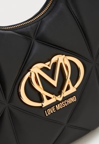 Musta tikkauksella varustettu nahkakäsilaukku, jossa on kultasävyinen sydän ja "M"-logo. Sujuva pinta, jossa on vetoketjukiinnitys ja erottuva geometrinen kuvio.