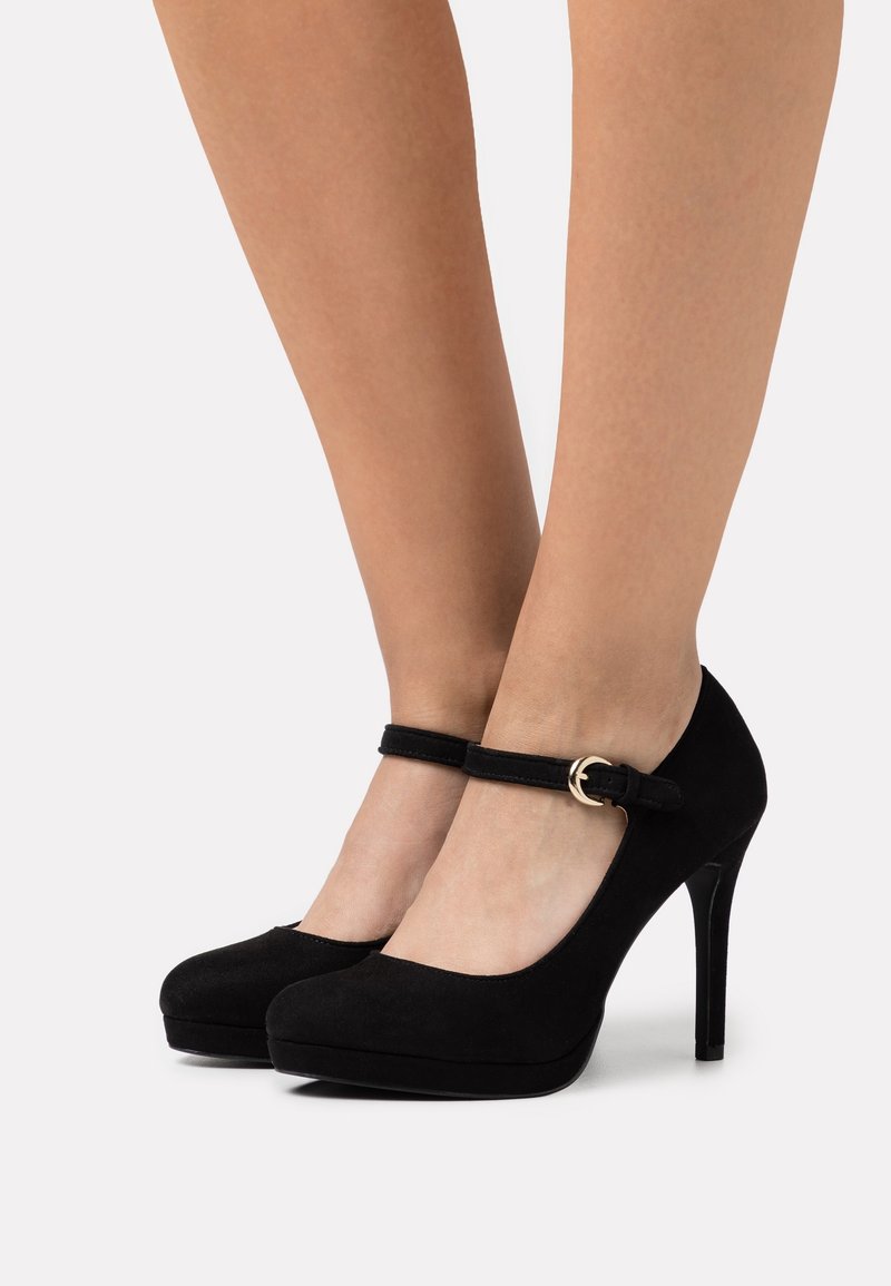 Chaussures Mary Jane à talons hauts en daim noir avec brides à la cheville et boucle dorée portées pieds nus sur fond blanc.