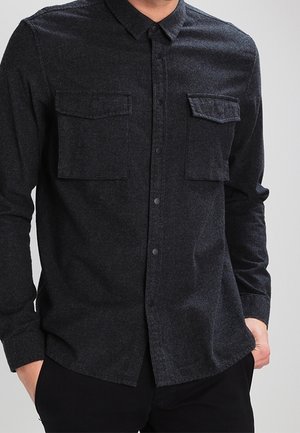 Camisa gris oscuro abotonada con dos bolsillos de solapa en el pecho, usada por una persona con las manos en los bolsillos de pantalones negros.