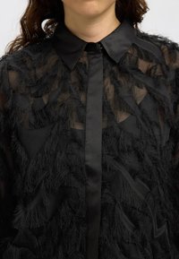 Black shirt features a sheer fabric with textured fringes and a classic collar, buttoned placket, and smooth satin accents.