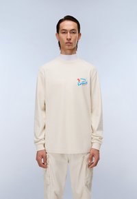 Κρέμα μπλουζάκι με μακριά μανίκια και στρογγυλή λαιμόκοψη, με μπλε και κόκκινο κείμενο στο αριστερό στήθος. Φοριέται με ταιριαστούς ανοιχτόχρωμους παντελόνες.