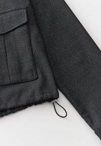 Manche de veste noire texturée avec poche à rabat et cordon ajustable à l'ourlet sur fond clair.
