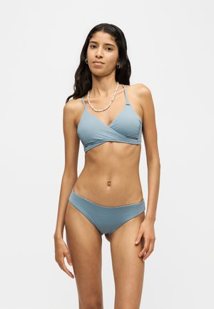 Jonge vrouw met lang donker haar die een lichtblauwe bikini en parelketting draagt, staand tegen een effen witte achtergrond.