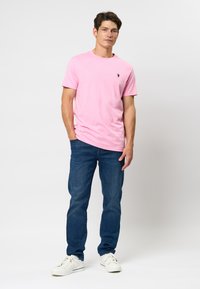 Man staat en draagt een lichtroze T-shirt met korte mouwen, blauwe jeans en witte sneakers tegen een effen witte achtergrond.