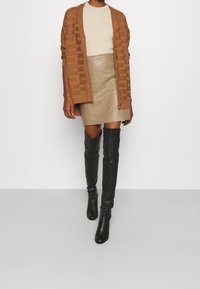 Cárdigan marrón texturizado sobre una blusa de canalé beige, combinado con una falda corta de cuero color marrón claro y botas negras hasta el muslo.