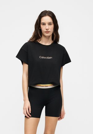 Femme portant un t-shirt court noir Calvin Klein et un short de cycliste noir avec ceinture Calvin Klein, debout devant un fond blanc uni.