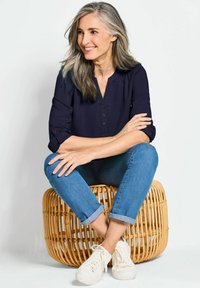 Navy Bluse mit Knopfleiste, kombiniert mit hellblauen aufgerollten Jeans und weißen Sneakers, sitzt auf einem geflochtenen Rattanstuhl.