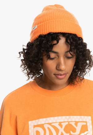 Mujer con cabello rizado que lleva un gorro de lana naranja y un sudadera naranja a juego con un logo blanco, mirando hacia abajo sobre un fondo blanco.