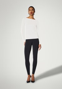 Weißes Langarmoberteil mit weit gefasstem Halsausschnitt, kombiniert mit schwarzen Leggings. Das Modell trägt schwarze High Heels, die eine moderne, taillierte Silhouette betonen.