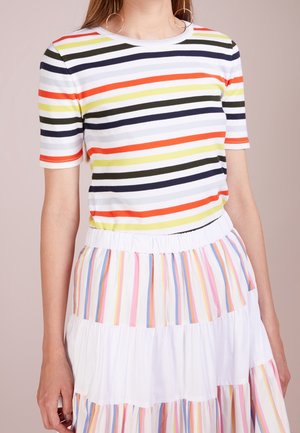 Kobieta w krótkim rękawie, w wielokolorową koszulkę w poziome paski oraz białą spódnicę z pionowymi pastelowymi paskami, stojąca na jednolitym tle.