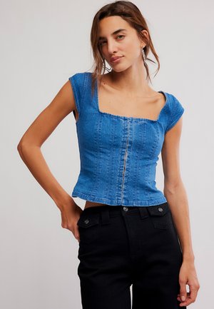 Νεαρή γυναίκα φορώντας εφαρμοστό μπλε τζιν κοντομάνικο τοπ με ραφές στο μπροστινό μέρος και μαύρο παντελόνι με ψηλή μέση, στέκεται μπροστά σε απλό φόντο.