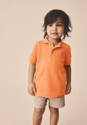 Junges Kind mit dunklen Haaren, das ein orangefarbenes kurzärmeliges Poloshirt und beige Shorts trägt, steht vor einem schlichten hellen Hintergrund.