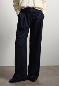 Marineblaue, weit geschnittene Hosen mit Falten, aus einem strukturierten Stoff gefertigt. Verfügt über Seitentaschen und einen taillierten Bund. Kombiniert mit schwarzem Schuhwerk.