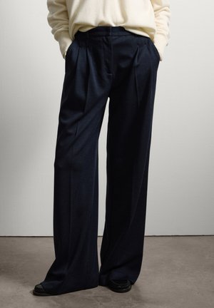 Pantaloni larghi navy con pieghe, realizzati in un tessuto strutturato. Presentano tasche laterali e una cintura su misura. Abbinati a calzature nere.