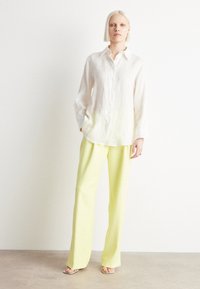 Lněná košile s dlouhým rukávem v barvě off-white, doplněná širokými kalhotami s vysokým pasem v světle žluté barvě. Model stojí na neutrálním pozadí.