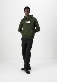 Hooded sweatshirt in olive green with a front kangaroo pocket and white printed logo. Black cargo pants and black sneakers complete the look.