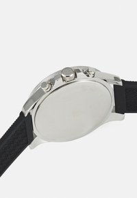 Silberner Edelstahl-Gehäuseboden mit strukturiertem Rand. Schwarzes Gummiarmband mit geometrischem Muster. Glatter, runder Gehäuseboden.