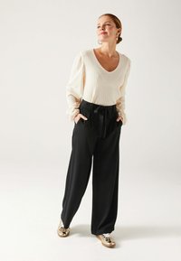 Pull côtelé crème avec un décolleté en V et des manches bouffantes, associé à un pantalon noir taille haute avec un nœud papillon et des poches latérales. Baskets dorées.