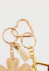 Nøglehænger med en gylden hjerteformet lås, et beige blomstermotiv, en tan frynse og en guldaccentueret vedhæng med en rød detalje.