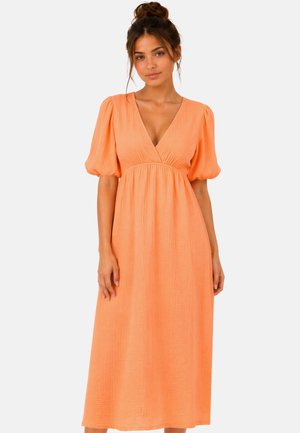 Vrouw met bruin haar in een losse knot, die een gestructureerde oranje midi-jurk draagt met pofmouwen en een diepe V-hals, staand tegen een effen achtergrond.