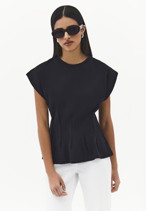 Kobieta z długimi ciemnymi włosami, nosząca okulary przeciwsłoneczne w oprawkach typu muszla żółwia, czarny bezrękawnik z wyściełanymi ramionami i białe spodnie na białym tle.