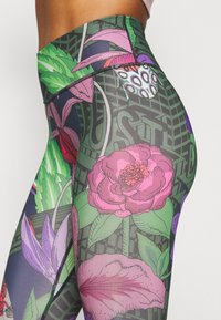 Gros plan sur une personne portant des leggings taille haute avec des motifs floraux et de feuilles colorés dans des tonalités de rose, vert, violet et noir.