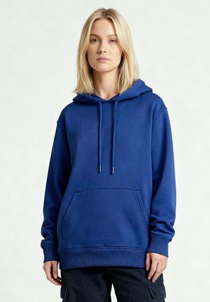 Jonge vrouw met stijl blond haar, gekleed in een blauwe hoodie met een zak aan de voorkant en zwarte cargobroek, staand tegen een effen achtergrond.
