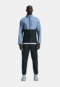 Hellblaue und schwarze Windjacke mit Reißverschluss, elastischen Bündchen und Taschen, kombiniert mit schwarzen Hosen und grauen Sportschuhen.