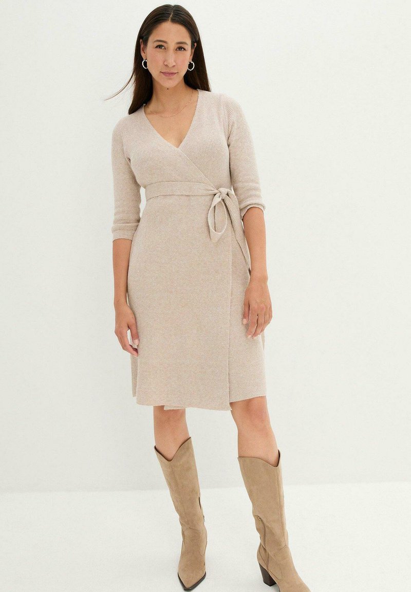 Abito avvolgente beige con scollatura a V profonda, maniche a tre quarti, tessuto testurizzato e vita con cintura. Abbinato a stivali alti fino al ginocchio color cipria.