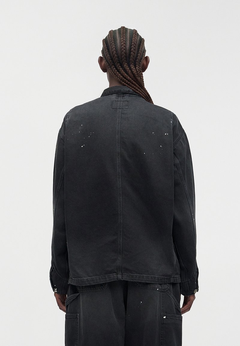 Person mit langen geflochtenen Haaren, die wegschaut, trägt eine dunkle, übergroße Jacke und Hose mit kleinen weißen Farbspritzern auf einfarbigem Hintergrund.