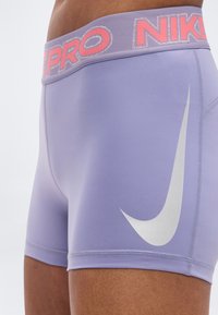 Γυναικεία σορτς Nike Pro σε ανοιχτό μωβ ύφασμα, με λευκό λογότυπο swoosh και ροζ ελαστική μέση με ανακλαστικές λεπτομέρειες.