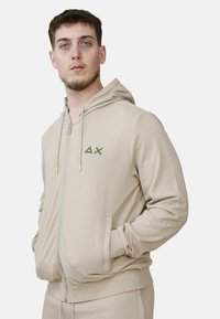 Felpa beige con zip, cappuccio, tasche a marsupio e logo ricamato verde. Tessuto morbido e polsini a coste per il massimo comfort.