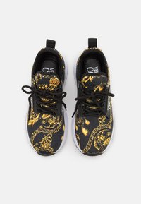 Svarta sportskor med guld barockmönster, med svarta snören, dragband och en vit sula. Den texturerade ovandelen ger visuellt intresse.