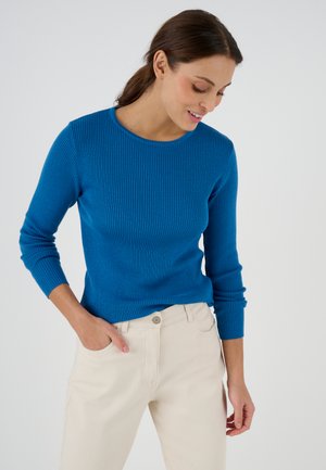 Vrouw met donker haar die een blauw geribd vest en lichtbeige broek draagt, kijkt naar beneden met één hand in de zak tegen een effen achtergrond.