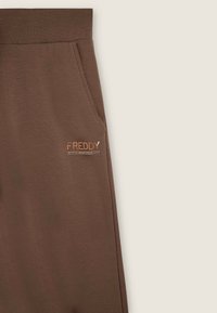Joggers marroni realizzati in morbido tessuto, dotati di tasche laterali e un piccolo logo metallico sul davanti. Tessuto liscio con una vestibilità comoda.