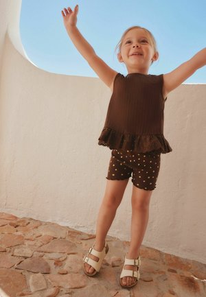 Junges Kind in braunem ärmellosem Oberteil und gepunkteten Shorts steht auf Steinboden mit erhobenen Armen vor weißer Wand und klarem blauem Himmel.