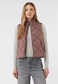 Gepolsterte Weste in Mauve mit hohem Kragen und Reißverschlusstaschen, getragen über einem hellgrauen Langarmshirt und kombiniert mit grauen Jeans.