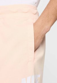 Calças de moletom rosa claro com um tecido texturizado, apresentando um bolso lateral e um detalhe em logotipo branco perto da bainha.