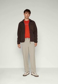 Donkerbruine fleece jas met een ritszak, een rode t-shirt eronder, lichtbeige broek en bruin-witte sneakers.