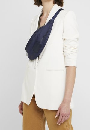 Person trägt ein weißes Sakko, beige Hose und eine dunkelblaue Umhängetasche mit sichtbarem Calvin Klein Jeans-Logo.