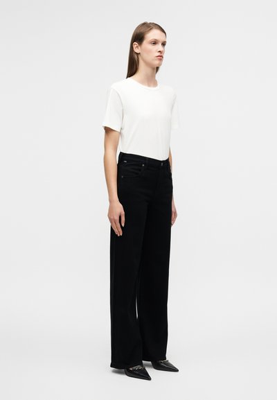 Λευκό κοντομάνικο μπλουζάκι συνδυασμένο με μαύρο παντελόνι φαρδύ στα πόδια και ψηλή μέση. Λείο ύφασμα με ελάχιστες ραφές· μαύρα παπούτσια με μυτερή μύτη.