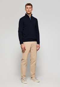 Suéter de punto azul marino con cremallera de cuarto de vuelta, combinado con chinos de color beige claro y zapatillas ligeras, que presentan un pequeño logo rojo en el suéter.