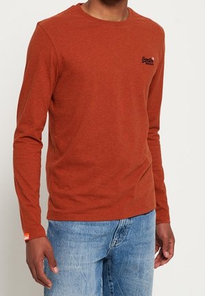 Mand iført rustfarvet langærmet skjorte med lille logo på brystet og blå denimjeans, stående mod en ensfarvet lys baggrund.