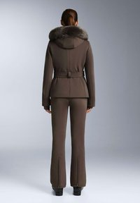 Hnědá voděodolná lyžařská bunda s límcem z umělé kožešiny, páskem v pase a přiléhavým střihem. Odpovídající hnědé lyžařské kalhoty se zipy na kotnících.