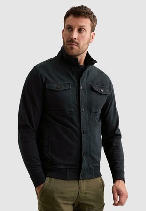 Man met kort haar die een zwart jack draagt met knoopsluitingen op de borstzakken en een groene broek, staand met handen gedeeltelijk in de zakken.