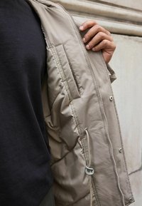 Jachetă bej, căptușită, cu închidere prin fermoar, buzunar lateral și șnur reglabil. Materialul are o textură fină și un finisaj mat.