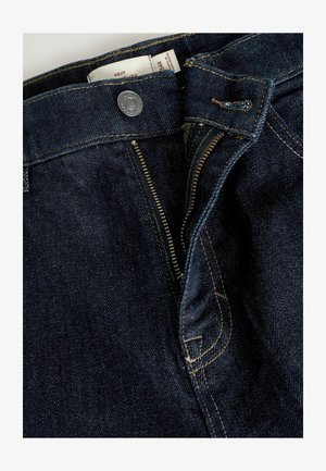 Tmavomodré džínsy z denimu so zipsovým zapínaním, kovovým gombíkom a kontrastným žltým prešívaním. Obsahujú štandardný dizajn s piatimi vreckami.