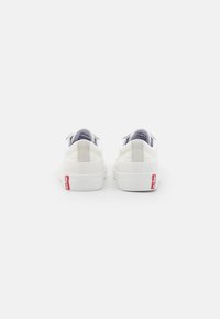 Baskets blanches avec une tige en cuir lisse, bout rond, semelle plate et un tag logo Levi's rouge au niveau du talon. Design minimaliste dans l'ensemble.