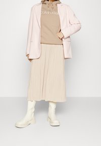 Béžová mikina Calvin Klein pod světle růžovým blejzrem, doplněná plisovanou béžovou sukní a bílými kozačkami nad kolena. Celkový vzhled s hladkými texturami.